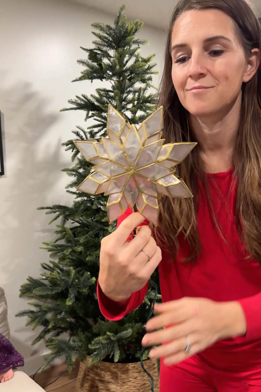 A woman dressed in red is holding a beautifully crafted stained glass star tree topper, examining it before placing it on top of the Christmas tree. The tree in the background awaits final touches.